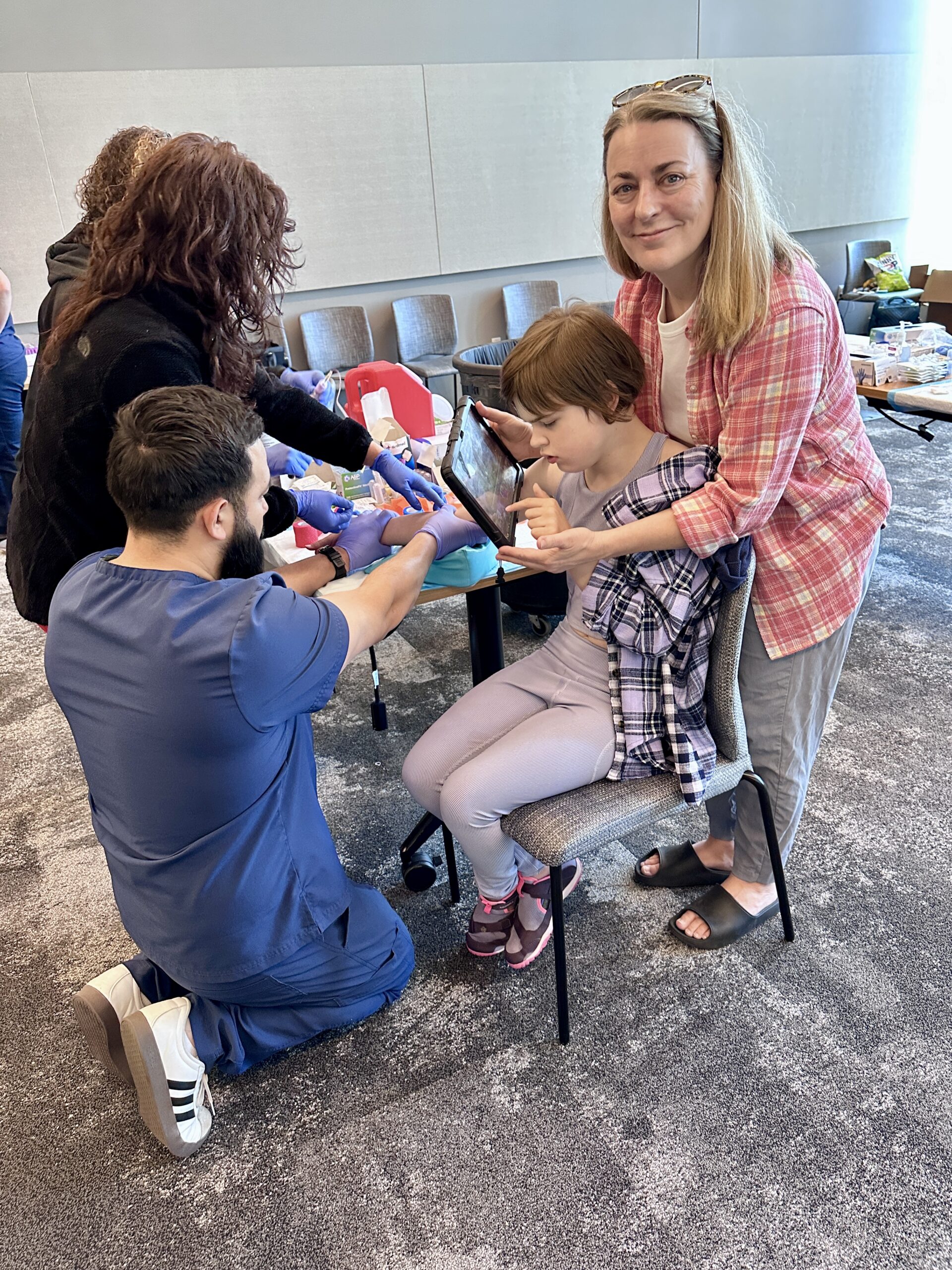 A child uses a tablet while a sample is collected at a KCNQ2 Biorepository event, with her mom beside her